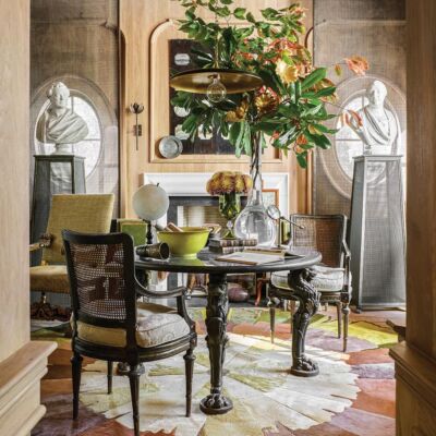 Table at center of home library designed by Barry Dixon. Dahlia rug on floor and vase of magnolia and other tree branches on table. Pair of busts on mesh pedestals in front of oval windows.