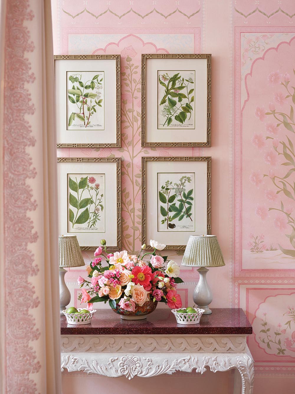 Console table with flower arrangement in pinks and corals under four botanical prints on pink, Indian-inspired wallpaper.