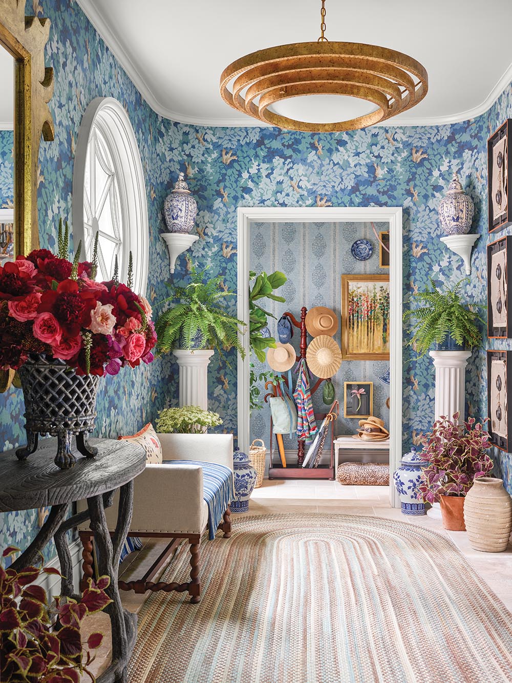 Hallway with blue floral tapestry wallpaper, demilune table with large arrangement of peonies, round window and round light fixture.