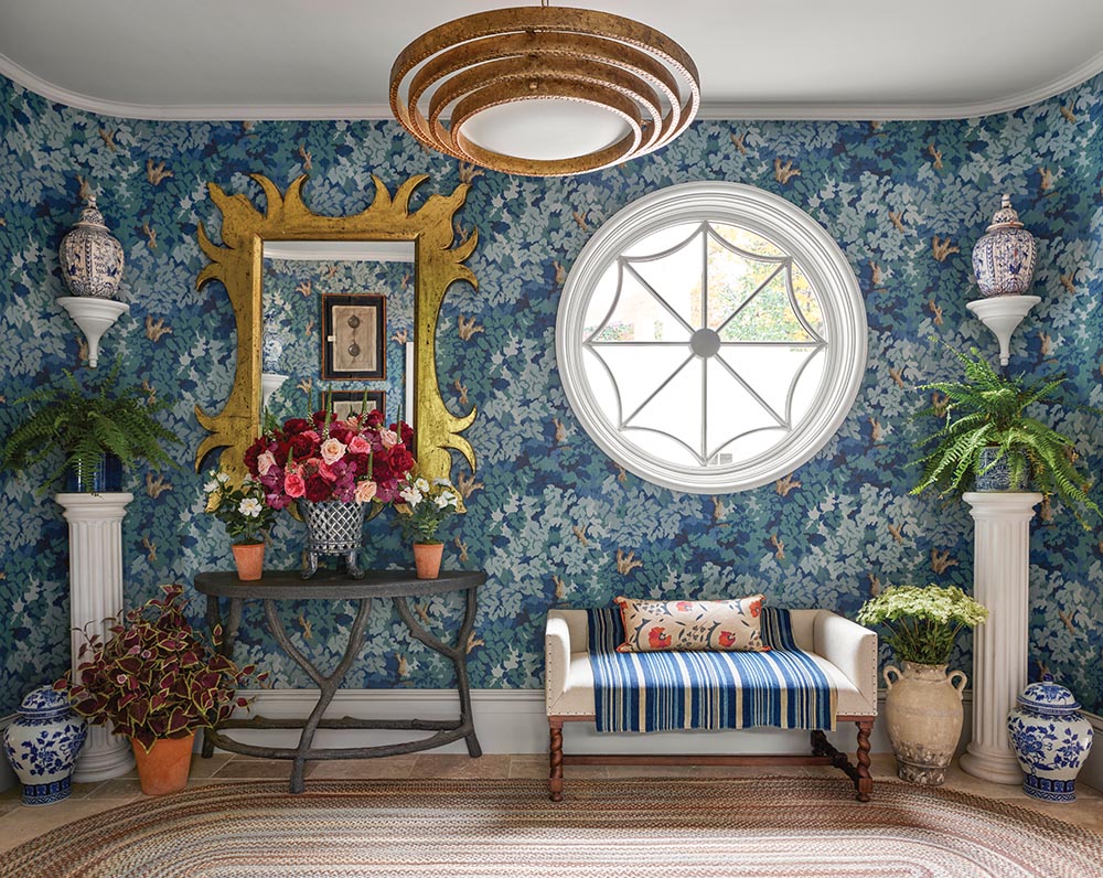 Gallery wall with blue floral tapestry wallpaper, large round window, gold-framed mirror, large arrangement of peonies on table, and ferns on column pedestals.