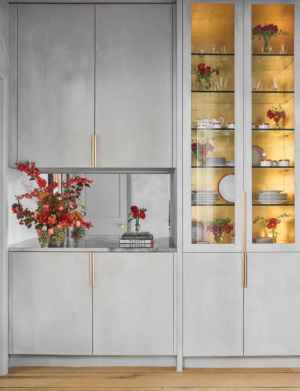 Cabinets witth a gray plaster pastellone finish from Domingue and counter with a red arrangement of pyracantha berries and flowers. Glass doored cabinet lined with gold wallpaper is filled with red and white china and vases of red ranunculus.