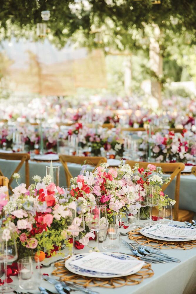 Red, white, and pale pink flowers sit nestled on a table with bright lime colored greenery. Blue patterned plates with menus on top sit next to the overflowing flowers.
