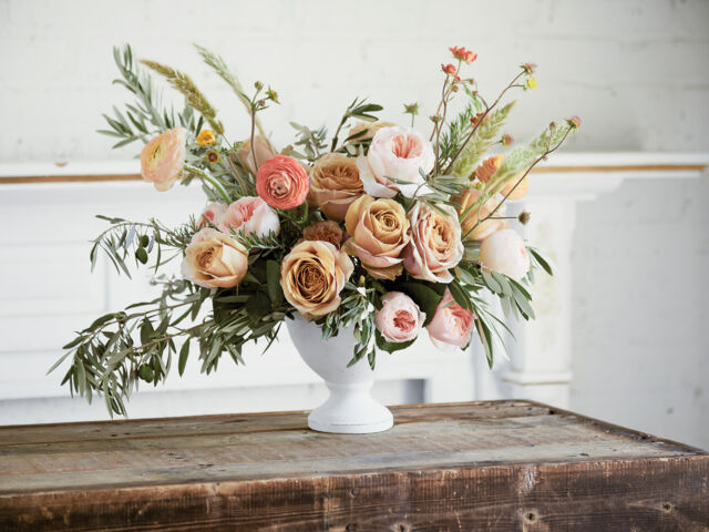 An arrangement of pink roses, poppy pods, olive branches, rosemary branches, wheat, and ranunculus sit in a white pedestal vase.
