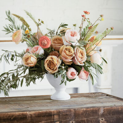An arrangement of pink roses, poppy pods, olive branches, rosemary branches, wheat, and ranunculus sit in a white pedestal vase.