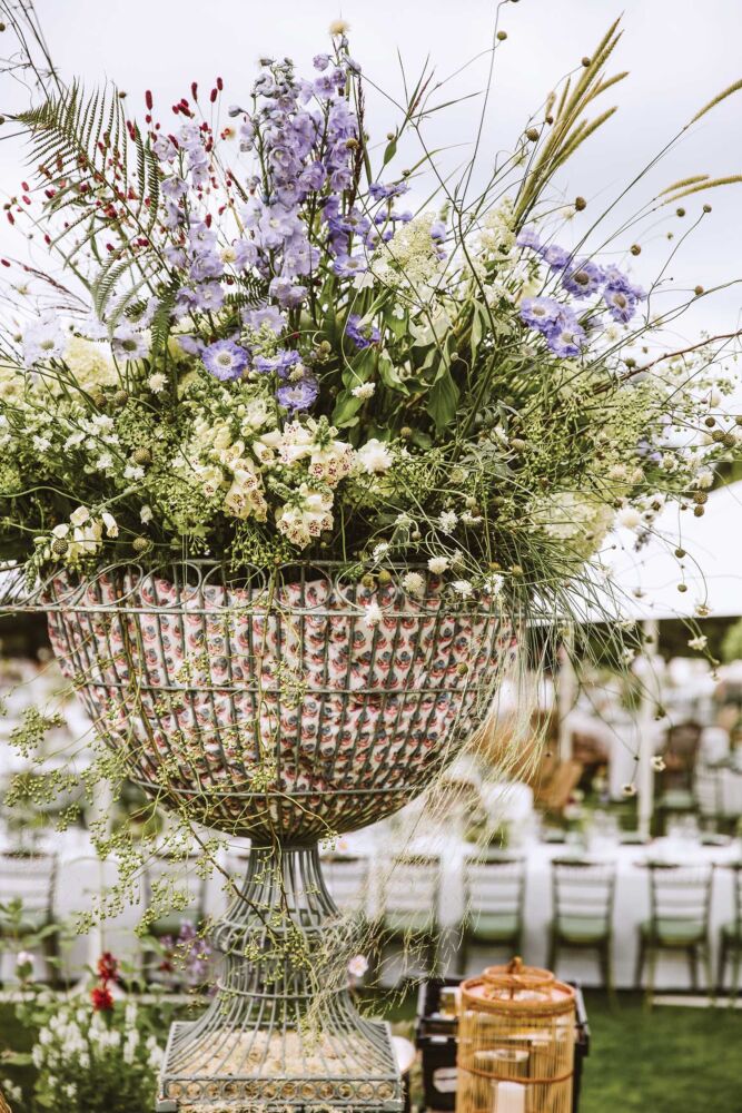 Large outdoor arrangement of scabious, foxgloves, delphinium, and Sanguisorba in a wire urn lined with block-printed fabric.
