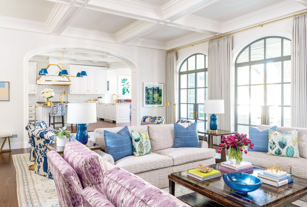 Purple and blue patterned furniture fill a white living room with sun peering in the large arched windows.