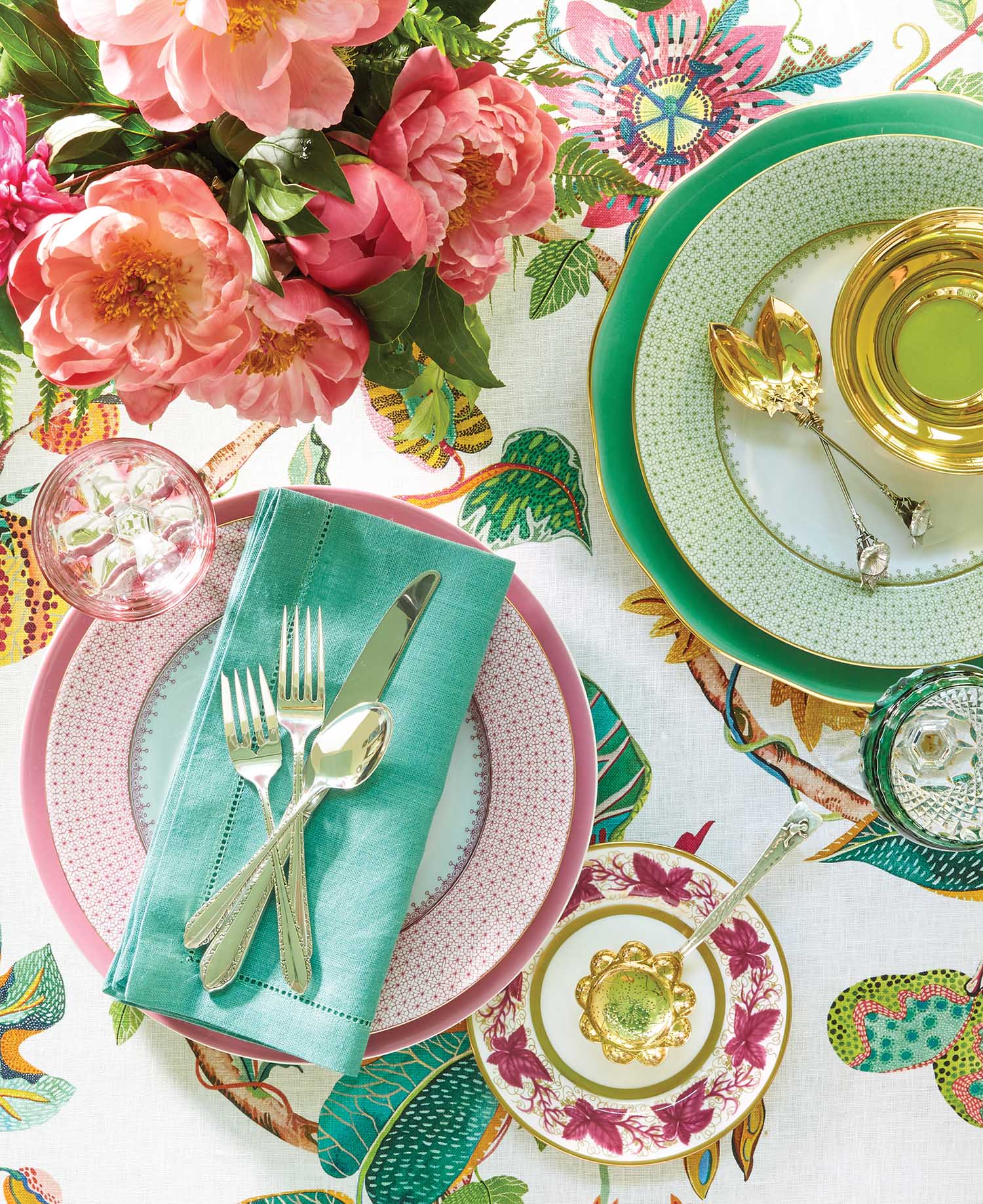 A turquoise napkin sits on top of a pink patterned plate with silverware. Pink peonies flow out of a vase next to a green patterned plate with gold silverware.