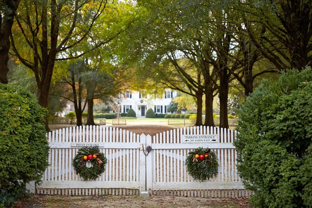 Two evergreen wreaths with fruit hang on a white picket fence.