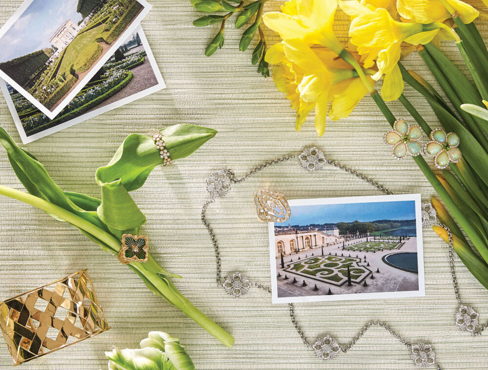 Diamond encrusted earrings, a gold ring, and a silver floral necklace lay next to pictures of Versailles.