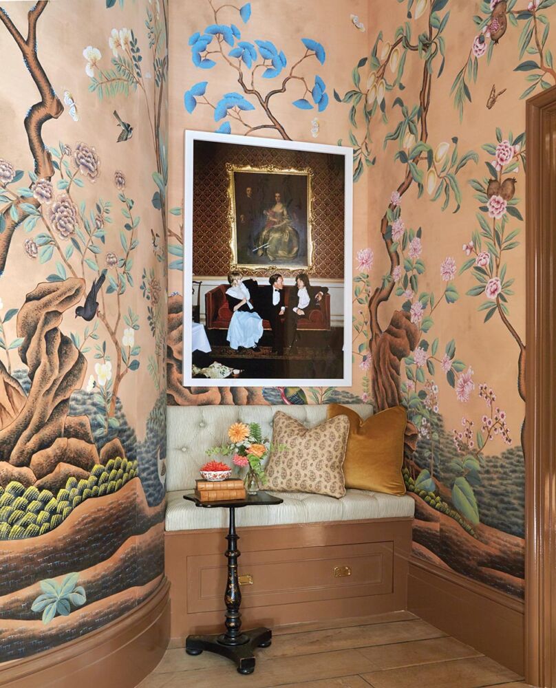 Built in bench in corner of gentleman's dressing room surrounded by scenic wallpaper. Slim Aarons photo of Mick Jagger, Marianne Faithful, and Desmond Guiness on wall. Small table with books, a vase of dahlias, and dahlia floating in bowl in front of bench.