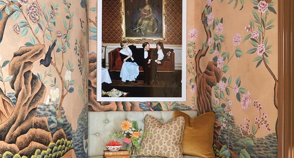 Built in bench in corner of gentleman's dressing room surrounded by scenic wallpaper. Slim Aarons photo of Mick Jagger, Marianne Faithful, and Desmond Guiness on wall. Small table with books, a vase of dahlias, and dahlia floating in bowl in front of bench.