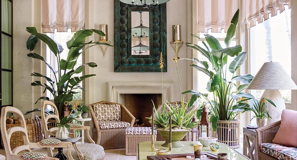 Screened porch with potted plants, wicker sofa and teak chairs flank an oversized coffee table. Wicker chairs sit in front of a fireplace. Designed by Ashley Whittaker for the Flower Atlanta Showhouse.