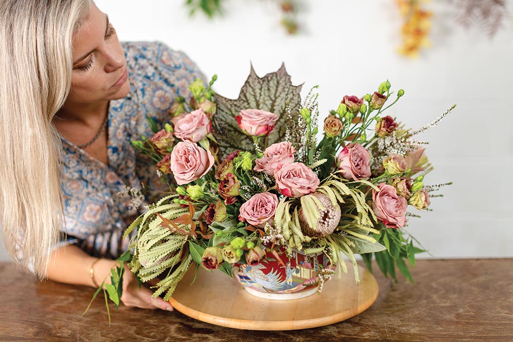 Holly Bryan adding Banksia, Lisianthus, and roses to flower arrangement in bowl.