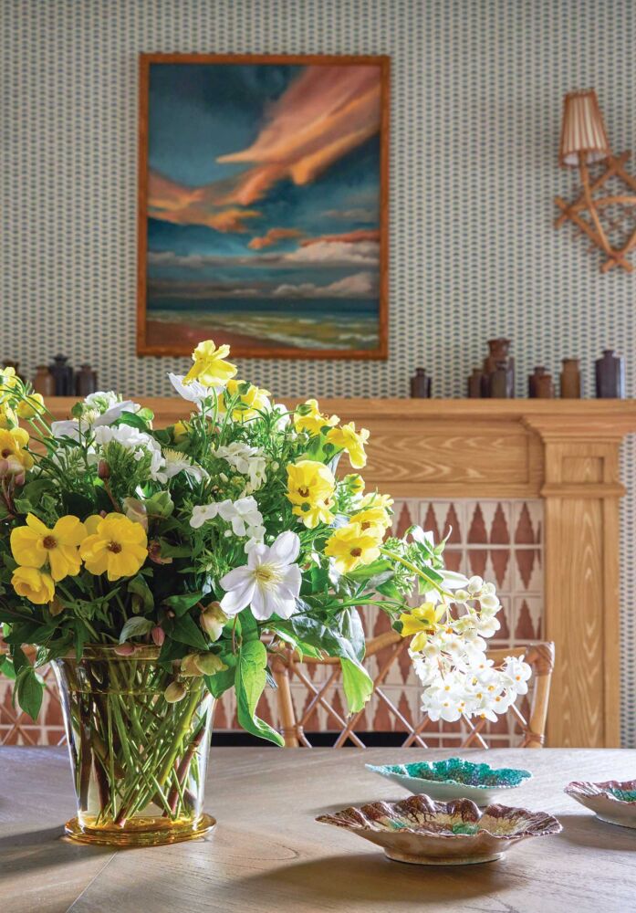 Arrangement of yellow and white flowers with pale lilac clematis on table in front of faux-painted mantel.