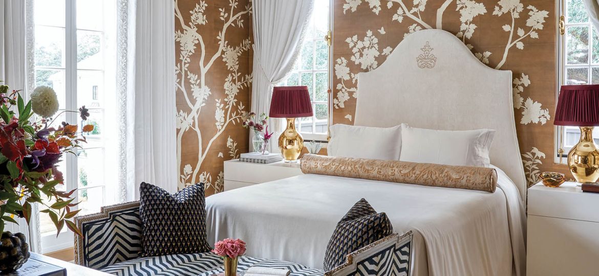 A bedroom has a cream bed and gold and white floral wallpaper.