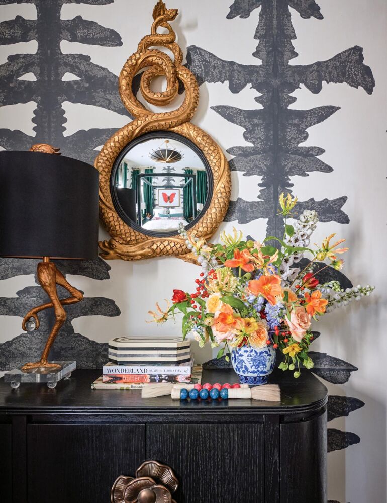 Black console topped with black-shaded brass bird lamp, books, box, flowers by Pick-A-Petal, with black and white graphic wallpaper and gold snake mirror on wall.