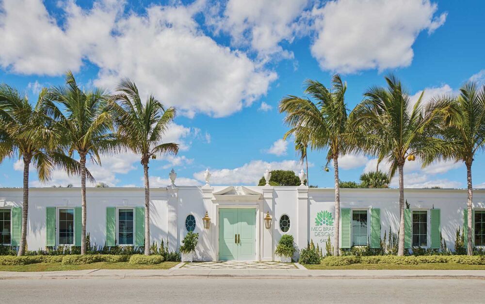 The exterior of Meg Braff Designs with pale green door and shutters, anchored by tall double doors, a broken pediment, brass carriage lights, and a line of palm trees.