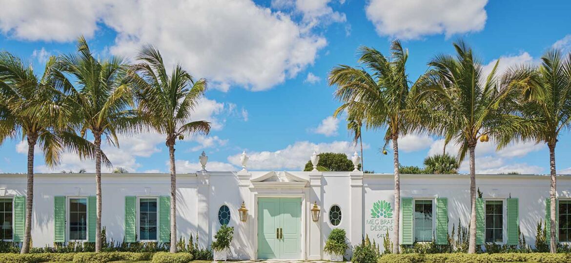 The exterior of Meg Braff Designs with pale green door and shutters, anchored by tall double doors, a broken pediment, brass carriage lights, and a line of palm trees.