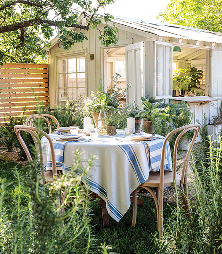 A centerpiece of potted plants on an alfresco table, nestled among rosemary in a California garden, beside a quaint white potting shed.