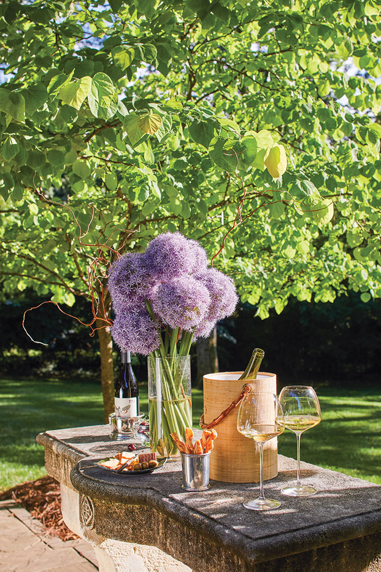 a vase of purple allium blooms wine, and appetizers on a stone outdoor table
