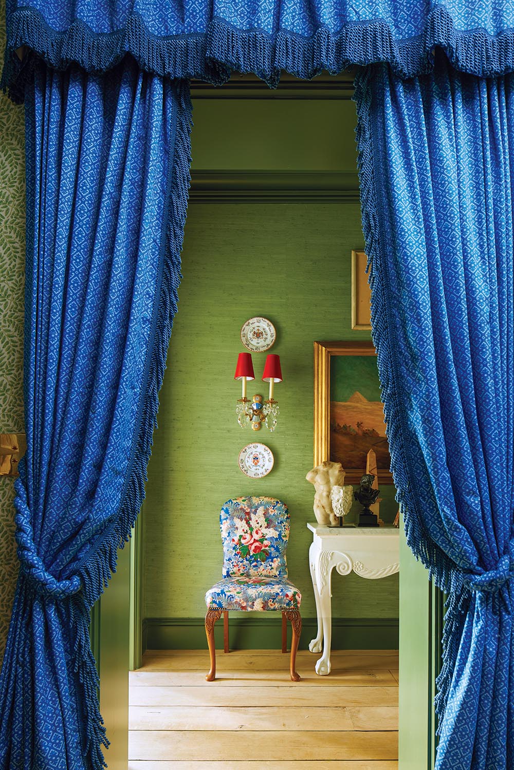 Blue portieres open onto bedroom hallway with chintz-covered chair under red-shaded sconce on the second floor of the Flower Atlanta Showhouse
