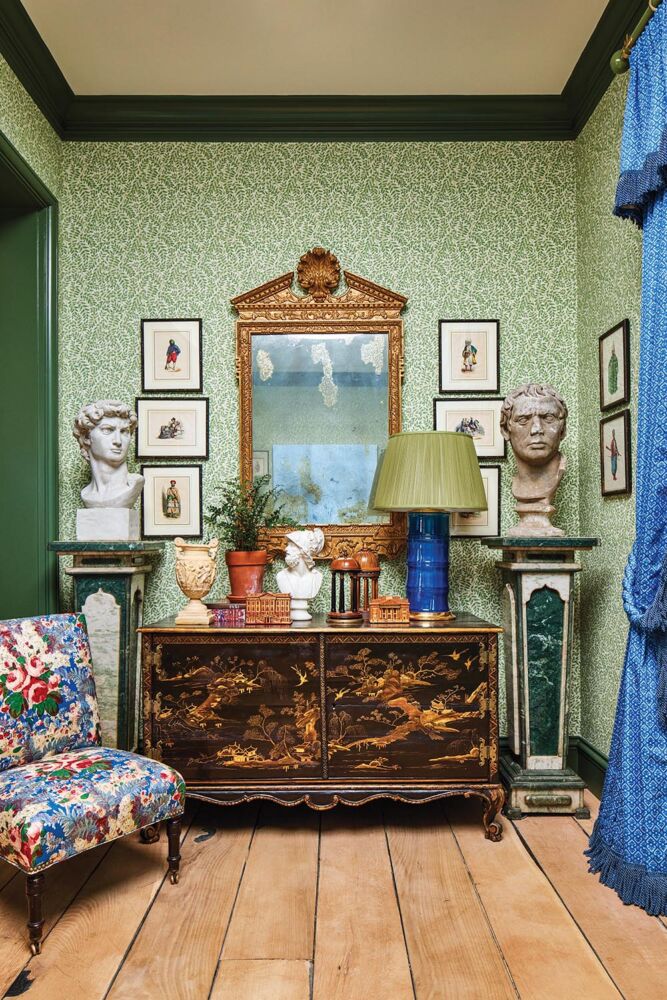 Antique chinoiserie chest flanked by busts on pedestals and topped with a blue lamp, architectural models, an urn and potted fern. Armless, chintz-covered chair in front of chest.