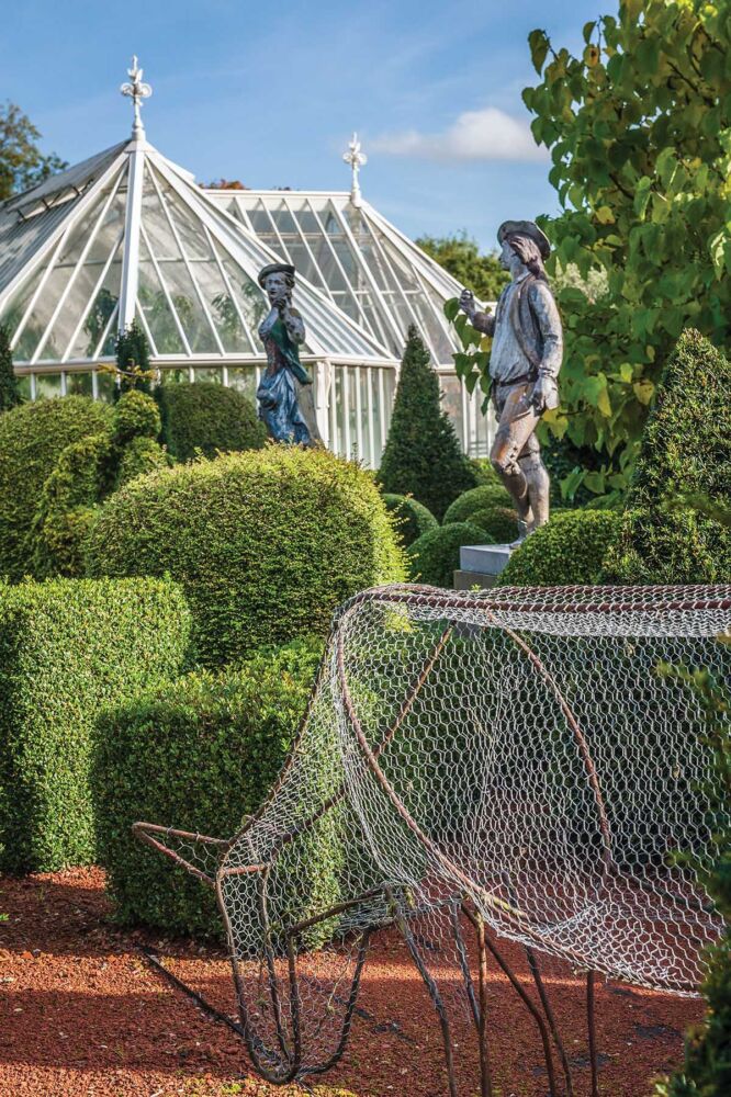 A wire-framed horse grazes at the edge of a topiary garden. Classical sculptures and the peaks of a glass house room stand above the topiaries.