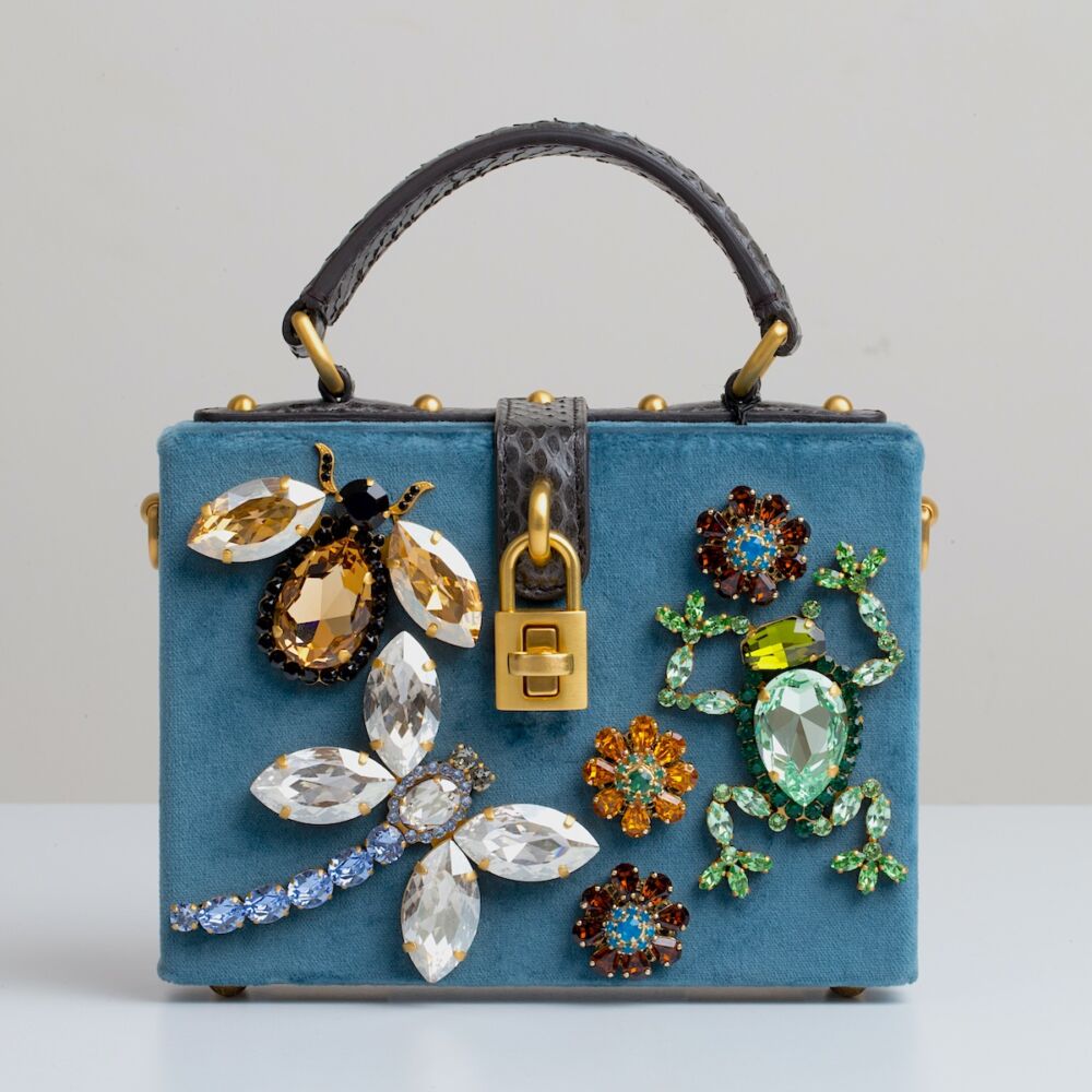 A bejeweled bee, a diamond encrusted dragonfly, and an emerald frog decorate a blue suede handbag.