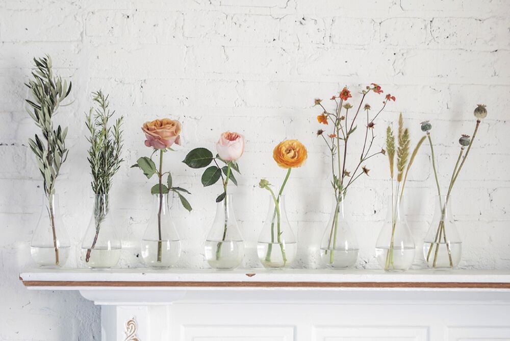 Clear glass vases with stems of olive branches, rosemary, Juliette roses, peach ranunculus, geum, wheat, and poppy seed pods.