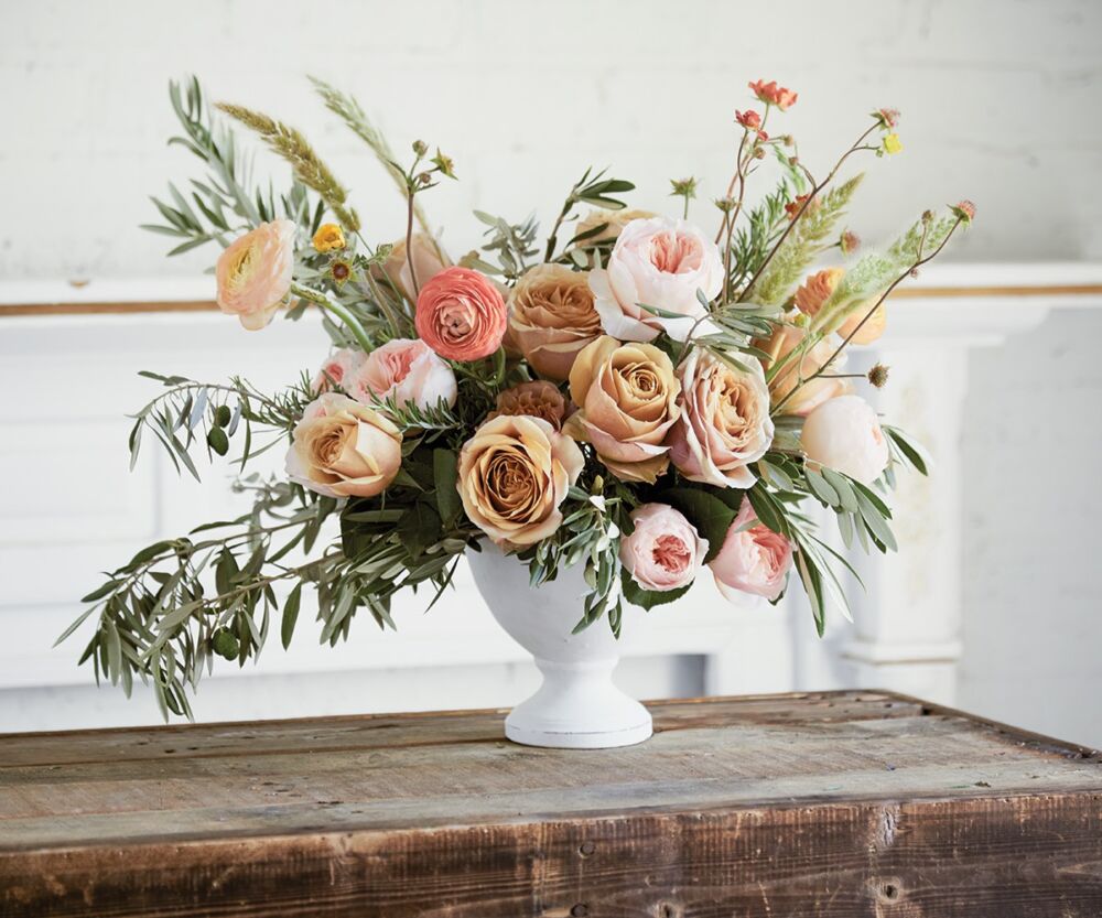 Arrangement of pink and gold roses and ranunculus in a white pedestal vase.