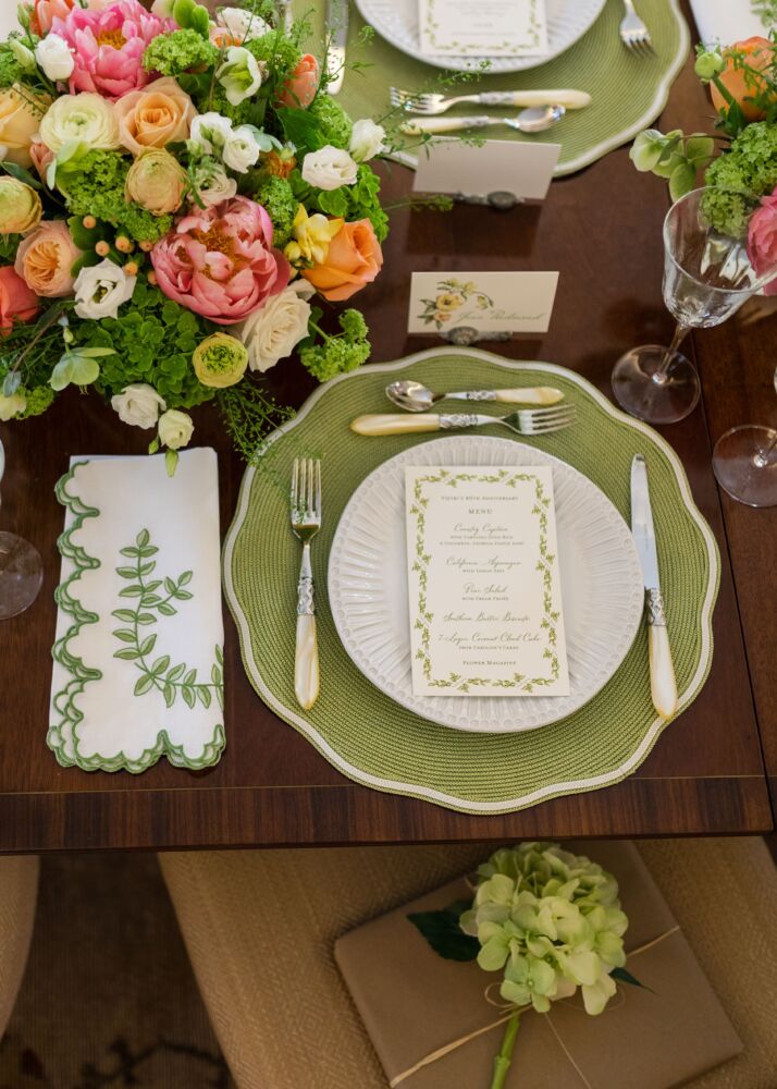A bouquet of greenery, and white, pink, and orange peonies and roses decorate a table with green placemats, green embroidered napkins, and white plates with a green bordered menu on top.