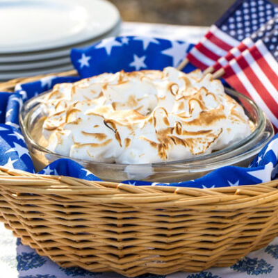 meringue-topped butterscotch pudding. The dish is wrapped in blue cloth with blue stars and nestled into a basket for a 4th of July meal. Two small American flags are also tucked into the basket