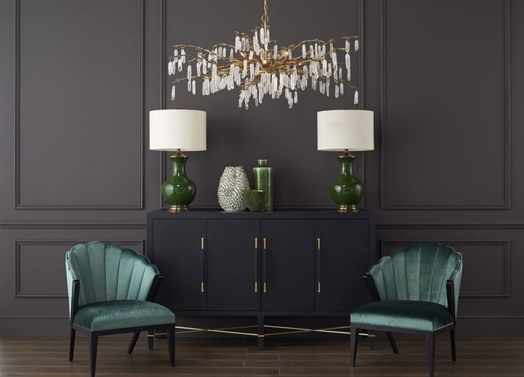 A botanically inspired chandelier design from Curry & Company, which features natural quartz crystals hanging from golden spokes that resemble branches of a tree, hangs in an elegant room painted charcoal gray. A pair of emerald green table lamps and vases stand on a sideboard with a black finish, which is flanked by a pair of curvy, green velvet side chairs.