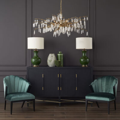 A botanically inspired chandelier design from Curry & Company, which features natural quartz crystals hanging from golden spokes that resemble branches of a tree, hangs in an elegant room painted charcoal gray. A pair of emerald green table lamps and vases stand on a sideboard with a black finish, which is flanked by a pair of curvy, green velvet side chairs.