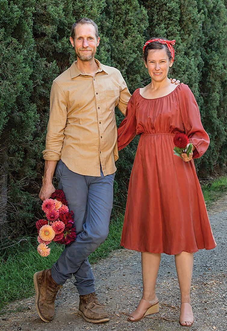Ceramics artist Josh Beckman of FBP Works and floral designer Holly Vesecky stand on a gravel road bordered with an allee of evergreens. Josh wears a tan button up, jeans and hiking books, and holds a bouquet of dahlias by his side; his other arm is around Holly. He crosses one foot casually over his standing leg. Holly wears a rust colored Bohemian-style dress, a scarf around her head and tan wedge sandals. She holds a red dahlia in one hand.