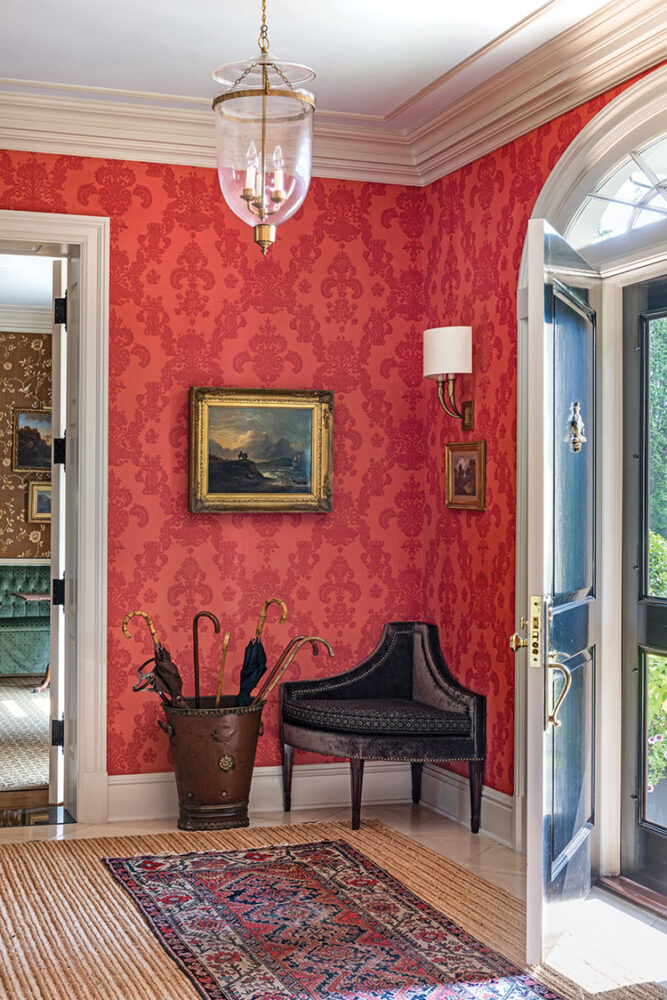 Red damask wallpapered entry hall with an oriental rug layered over a larger sea grass rug in a home designed by Matthew Patrick Smyth. A formal antique corner chair, traditional moldings, and gold framed antique paintings finish the space.