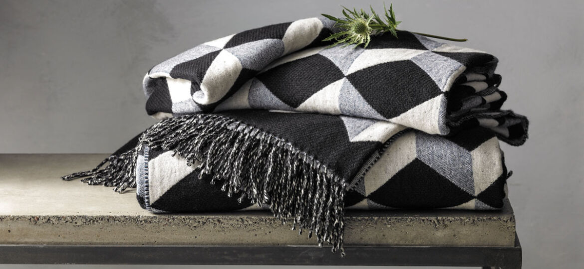 menswear-inspired wool and cashmere throw in a geometric patter of black, gray, and off-white, folded and placed on a bench