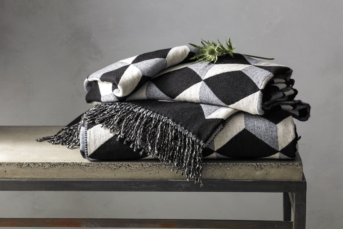 menswear-inspired wool and cashmere throw in a geometric patter of black, gray, and off-white, folded and placed on a bench
