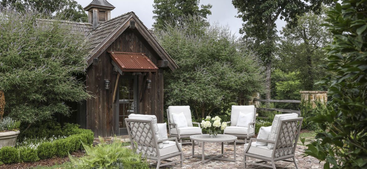 A set of 4 white-cushioned patio chairs, available through Blackjack Gardens in Birmingham AL, surround a small white table on a brick patio. The patio is surrounded by a lush green garden. The adjacent home is a rustic cabin with an aged metal overhang and a classic weathervane on roof.
