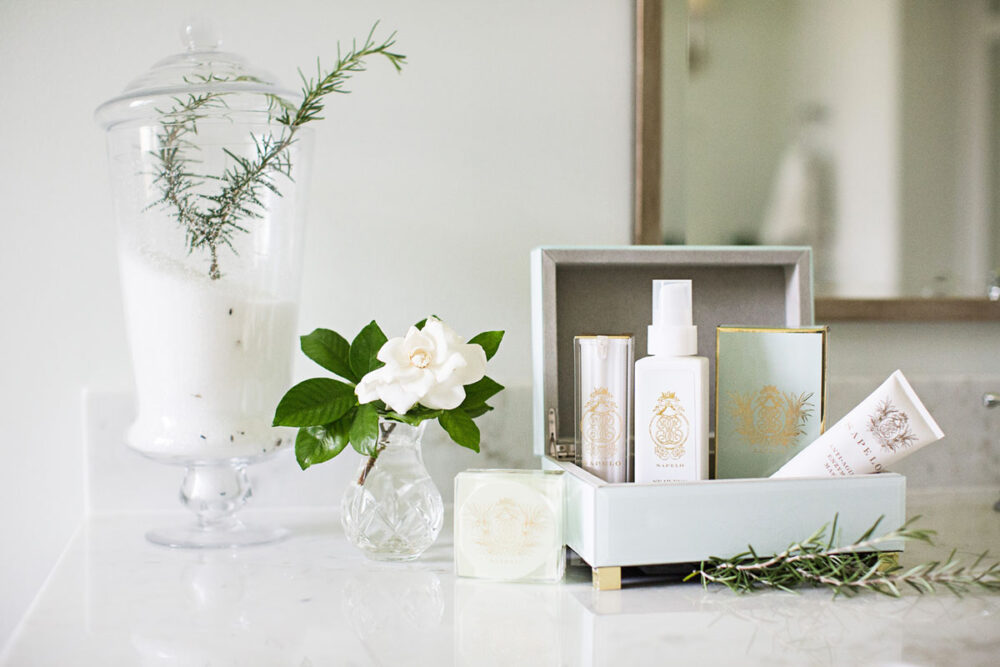 A white bathroom countertop, decorated with a glass container filled with bath salts and rosemary, a gardenia bloom in a bud vase, and Sapelo products in a pale green box