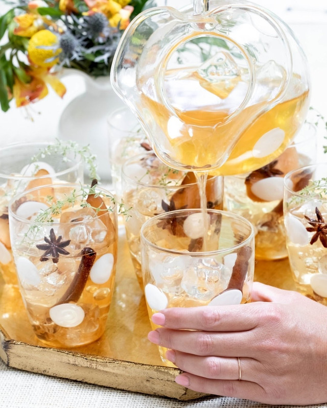 A glass pitcher and tumblers filled with a chilled cider, with cinnamon sticks and star anise, set on a wooden tray painted gold