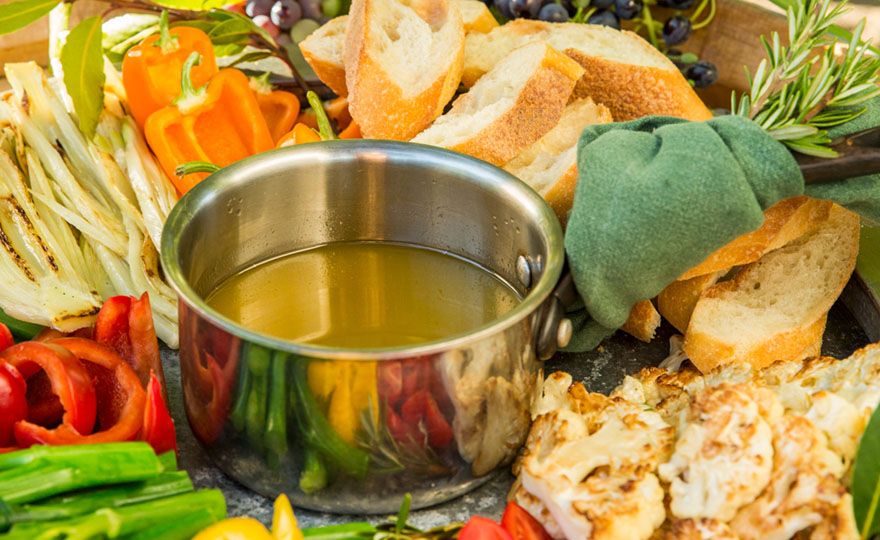 A silver dish of bagna cauda on a platter of sliced colorful sweet peppers, tomatoes, assorted breads for dipping, and grapes