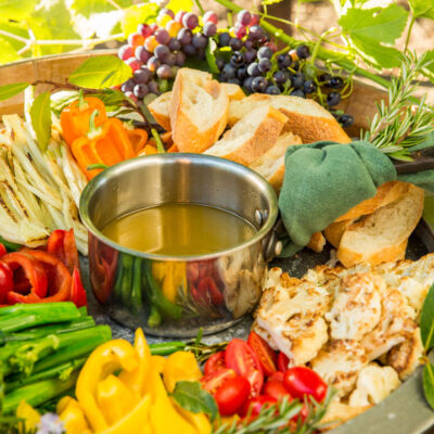 A silver dish of bagna cauda on a platter of sliced colorful sweet peppers, tomatoes, assorted breads for dipping, and grapes