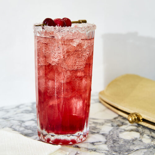 Highball glass filled with ice and Cranberry Shrub Cocktail from Very Merry Cocktails by Jessica Strand. The glass is rimmed in sugar and garnished with a skewer of cranberries. A gold clutch lays on the gray and white marble counter beside it.
