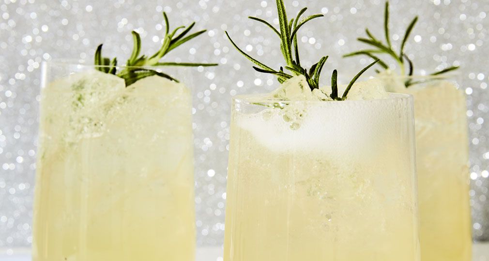 Three glass tumblers filled with Evergreen Sparkler, a fizzy lemon, rosemary, and gin cocktail from the book Merry Cocktails by Jessica Strand. Rosemary sprigs garnish each glass. The background is a glittery silver.
