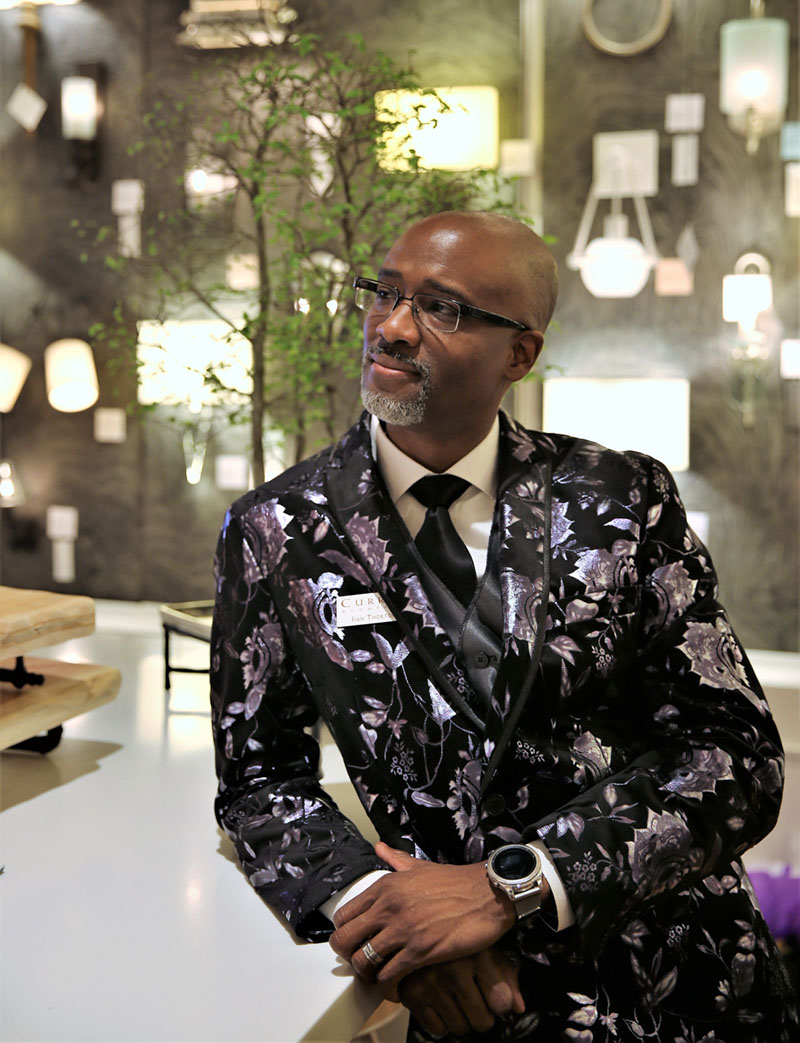Currey and Company Senior Lighting Designer Ian Thornton, creator of the Multi Drop Pendant Collection, stands in the showroom wearing a dark floral print suit coat.