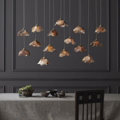 A light fixture of 14-pendant lights, with flower-shaped shades made from shells, hangs over a dining room table against a backdrop of wainscoting painted charcoal gray. The fixture is part of Currey and Company’s new Made to Measure Pendant Collection