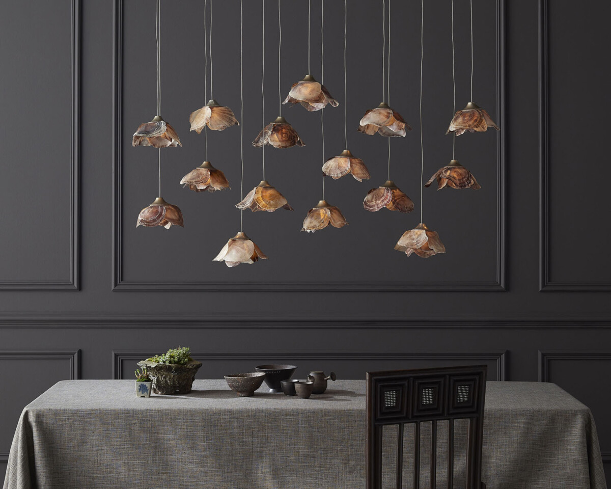 A light fixture of 14-pendant lights, with flower-shaped shades made from shells, hangs over a dining room table against a backdrop of wainscoting painted charcoal gray. The fixture is part of Currey and Company’s new Made to Measure Pendant Collection