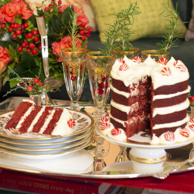 A holiday dessert table featuring a 4-layer red velvet cake on a pedestal and champagne in glasses garnished with rosemary sprigs and cranberries. In the background, an arrangement of red flowers fills a silver basket.