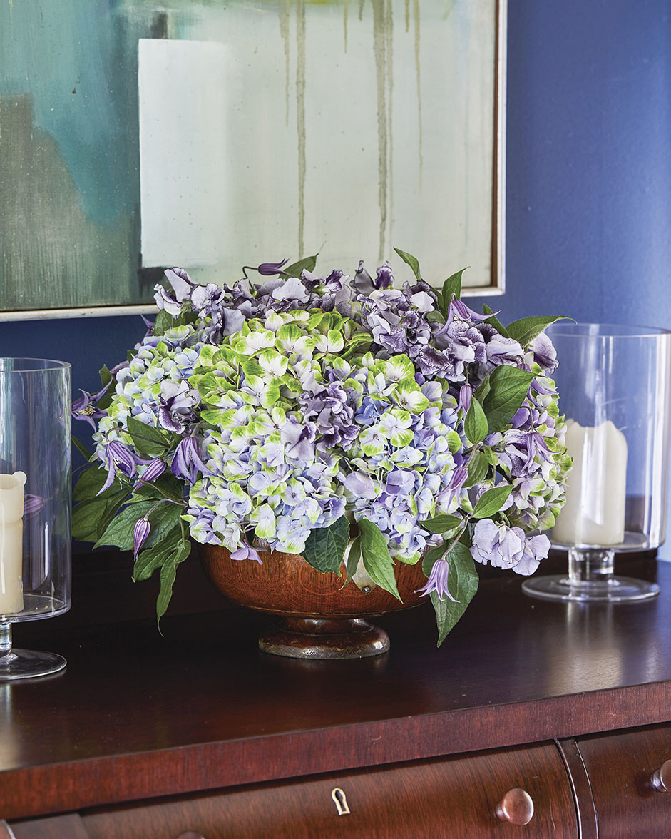 A purple floral arrangement by Kirk Whitfield of K & Co Flowers in a room painted royal blue. The arrangement, which includes sweet peas, hydrangea, and clematis, fills a large bowl set on a dark wood sideboard. An abstract painting, also featuring blue tones, hangs in the background